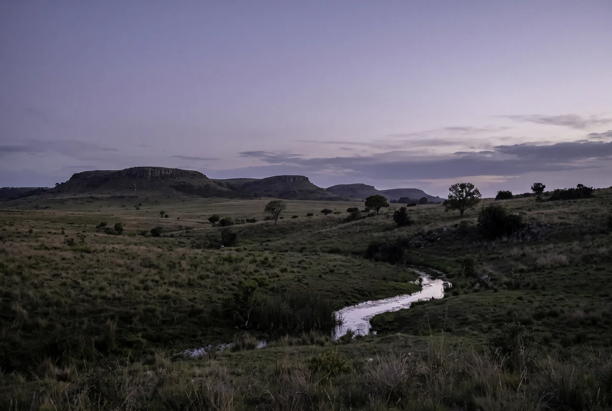 Low flat-topped hills and grassland with a small stream under a lavender sky.