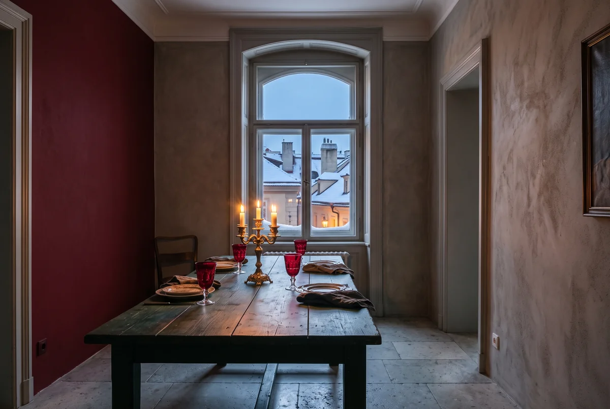 A garnet-red wall, grey plaster, a dark green table, red glass goblets and candles by a snowy window.