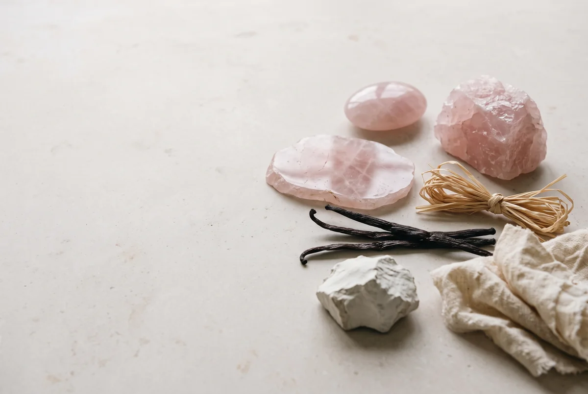 Raw and polished rose quartz, raffia, vanilla pods, white clay and raw silk on pale stone.