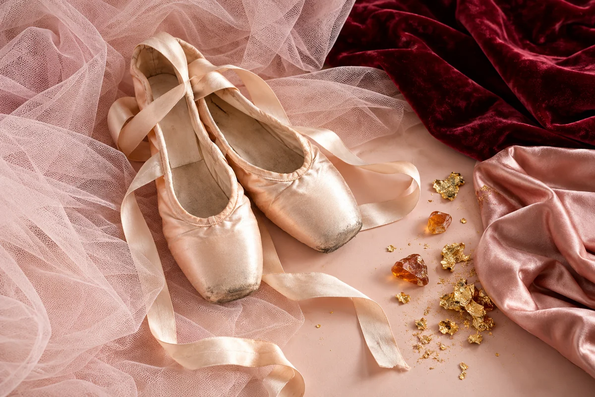 Blush tulle and pointe shoes lie beside satin ribbon, rosin, gold leaf and deep red velvet.