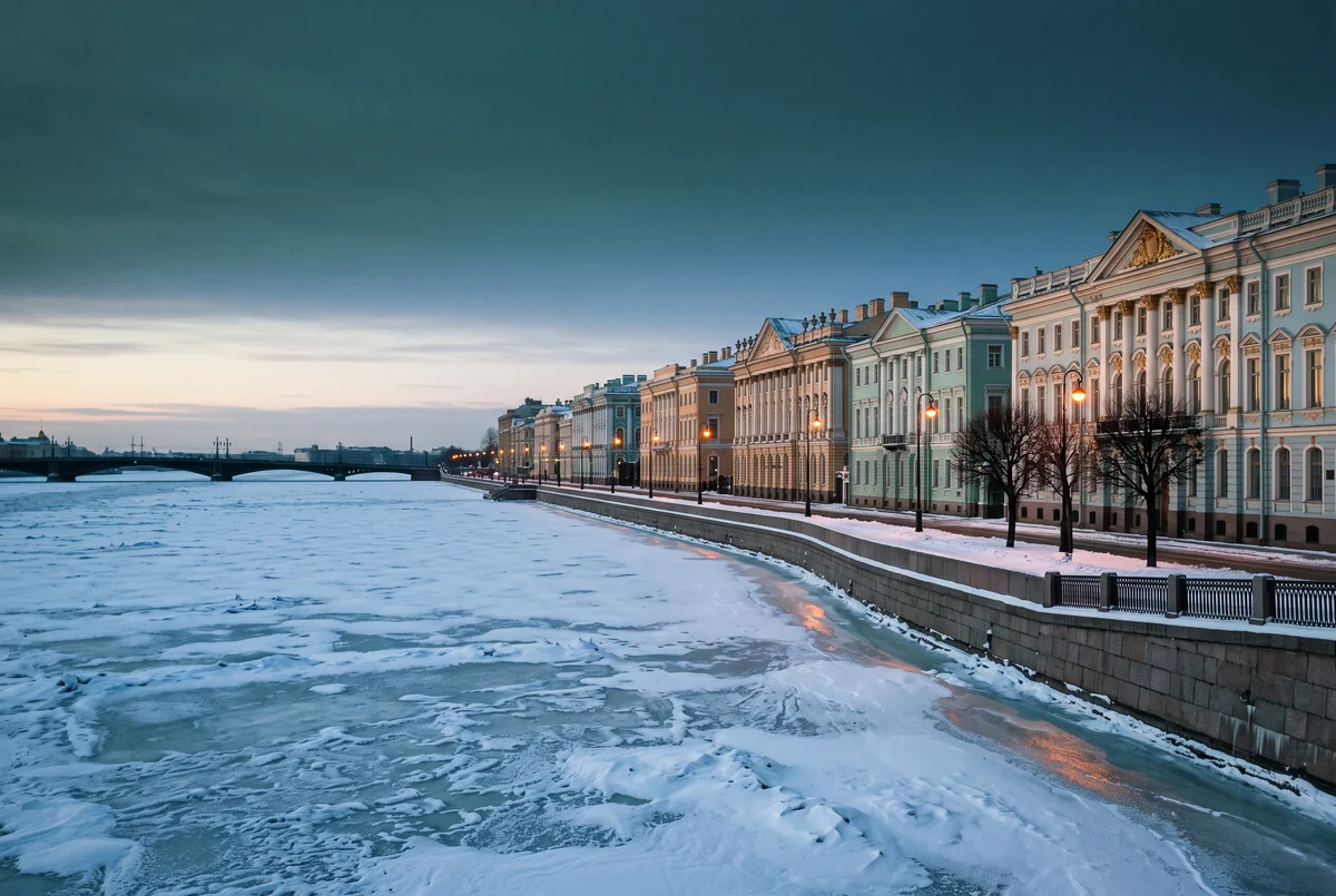 Snow on a frozen river beside a long embankment of pale classical facades with lamps lit.